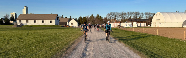 A group of cyclists riding on a gravel road through some farmer's fields. Two tall buildings poke up over a barn on the left side.