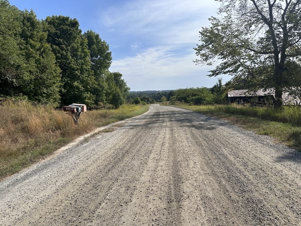A gravel road, stretching to the horizon. Green trees line the left side of the road, while a farm building is visible on the other side.